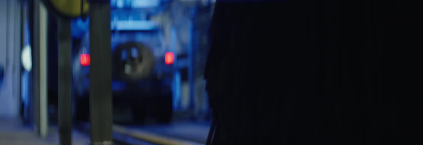 A mudspattered suv with a rearmounted spare tire moves slowly through a dark car wash tunnel its red taillights glowing vividly beneath the intense blue industrial lighting as it passes the cleaning brushes