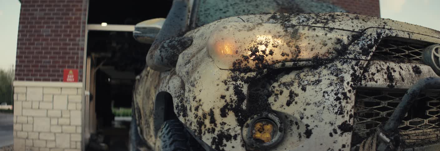 The front of a white toyota 4runner is shown covered in thick dark mud splatters across the grille and bull barthe camera pans across the dirty vehicle at a car wash location under the soft light of a setting sun