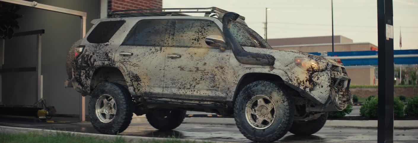 A heavily mudsplattered suv slowly exits an automated car wash bay into the daylight the vehicle is equipped with offroad gear and a roof rack showing the scale of its recent adventure