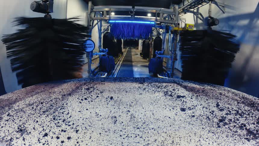 A dirty car hood moves through an automated wash tunnel where large black and blue brushes spin under bright lights to clean off thick mud and debris from the surface of the vehicle