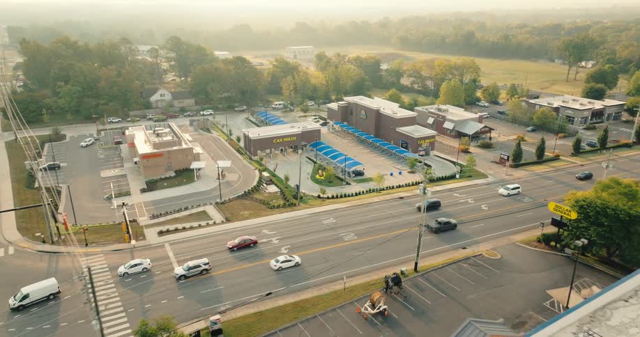 An aerial drone shot pulls back from a brick car wash with blue canopies to reveal a busy four way intersection filled with cars and surrounding businesses under a hazy morning sky