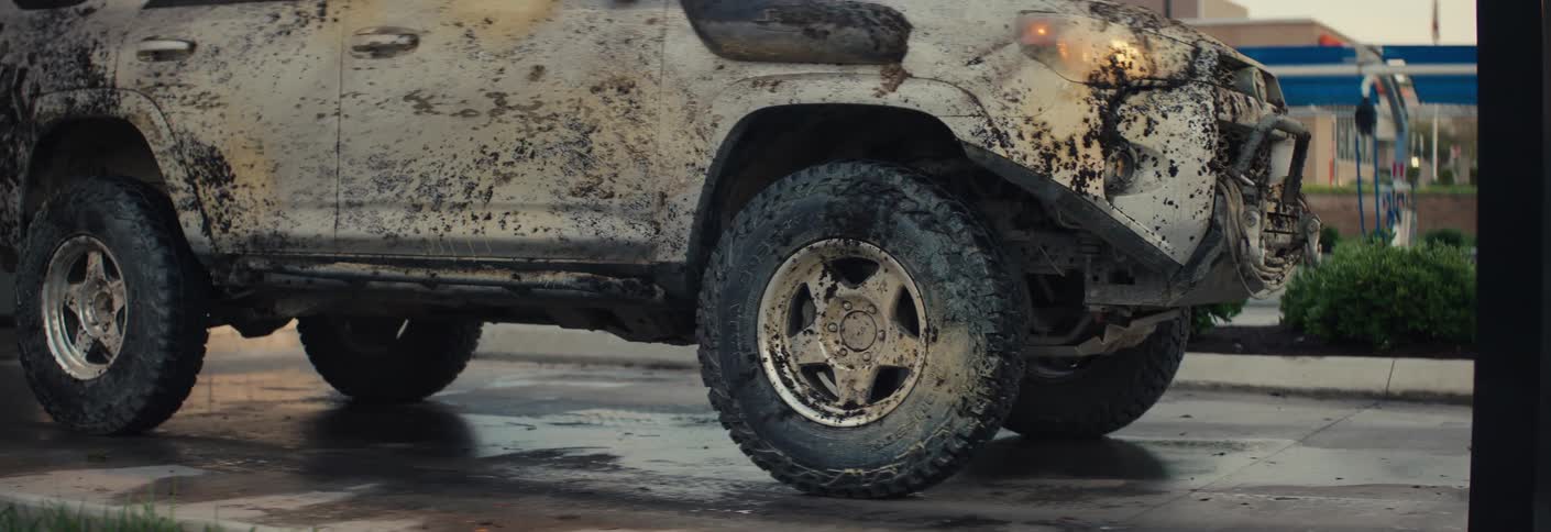 A mudcaked suv slowly exits a car wash bay its tires and white body panels heavily layered in thick dirt that contrasts against the clean wet pavement