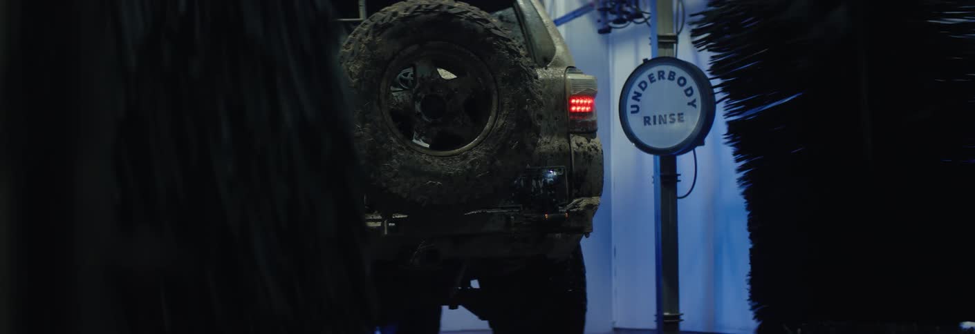 A mudcovered suv slowly moves through an automated car wash passing large rotating brushes the scene is dimly lit with blue tones highlighting an underbody rinse sign as the vehicle is cleaned