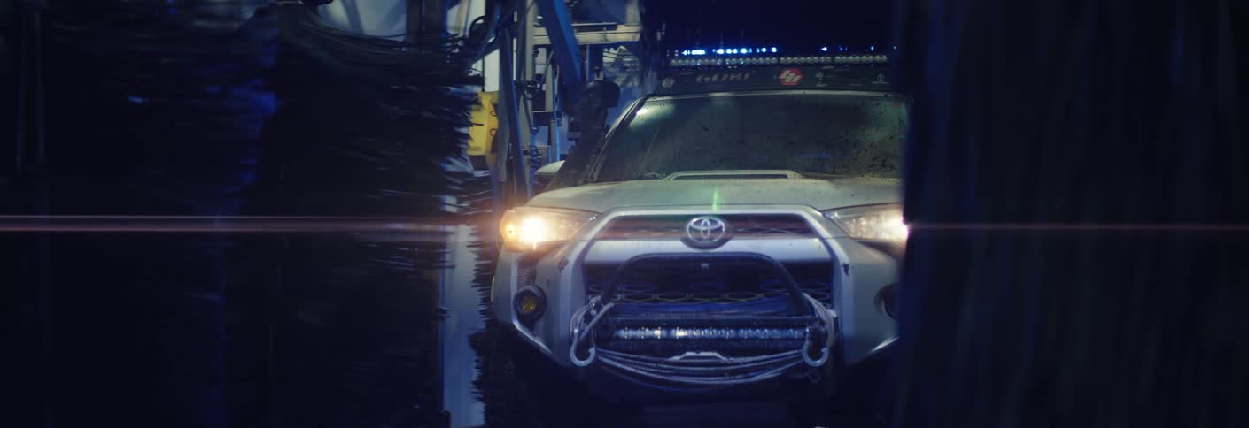A mudcovered toyota suv with its headlights on slowly enters an automated car wash bay large spinning brushes activate on the sides as the rugged vehicle prepares for a cleaning