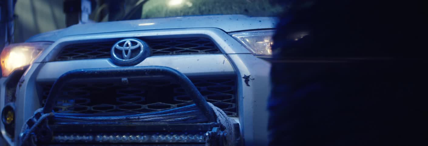 A white toyota suv drives into a car wash tunnel its headlights casting a bright lens flare as massive cleaning brushes spin against the front bumper under vibrant blue industrial lighting