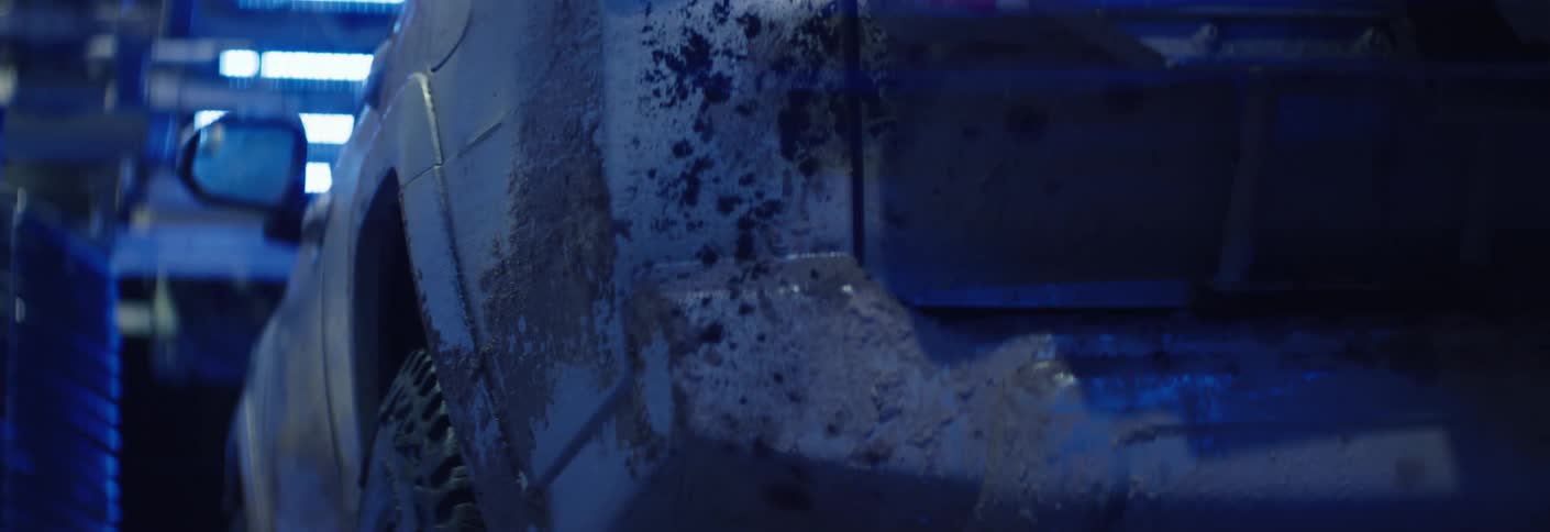 A mud caked white suv with an off road vehicle moves into a dimly lit car wash bay under blue lighting
