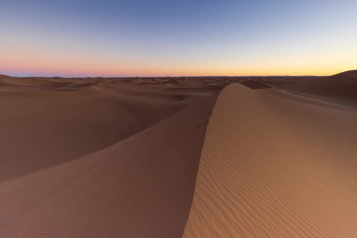 A view of a vast desert shows ripples on the sand dunes as the sun sets and the sky fades from orange to dark blue