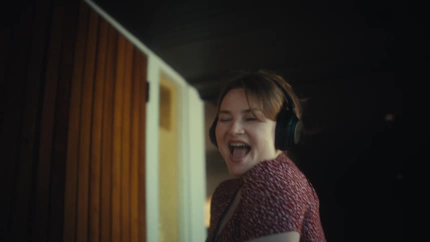 A woman joyfully dancing and singing into her phone while walking through a dark hallway toward a brightly lit home