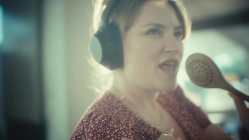 A young woman wearing headphones sings into a wooden spoon while dancing in a sunlit room