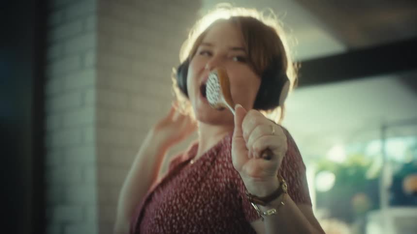 A young woman wearing large black headphones sings and dances enthusiastically in a sunlit room while holding a wooden spoon like a microphone