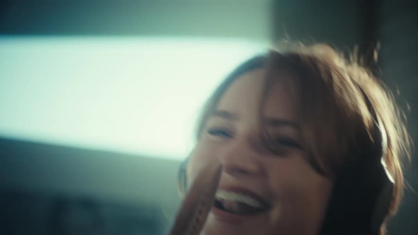 A young woman wearing large headphones sings loudly and dances while using a wooden spoon as a microphone in a kitchen
