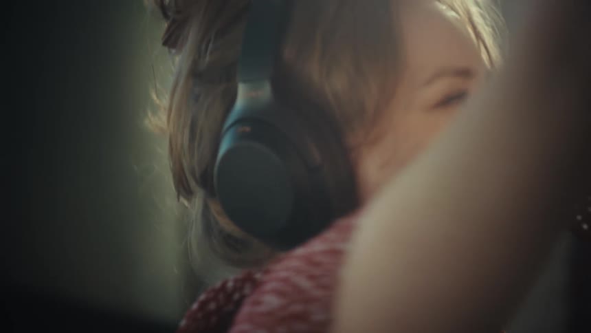 A young woman wearing large headphones sings loudly while using a wooden spoon as a microphone in a kitchen