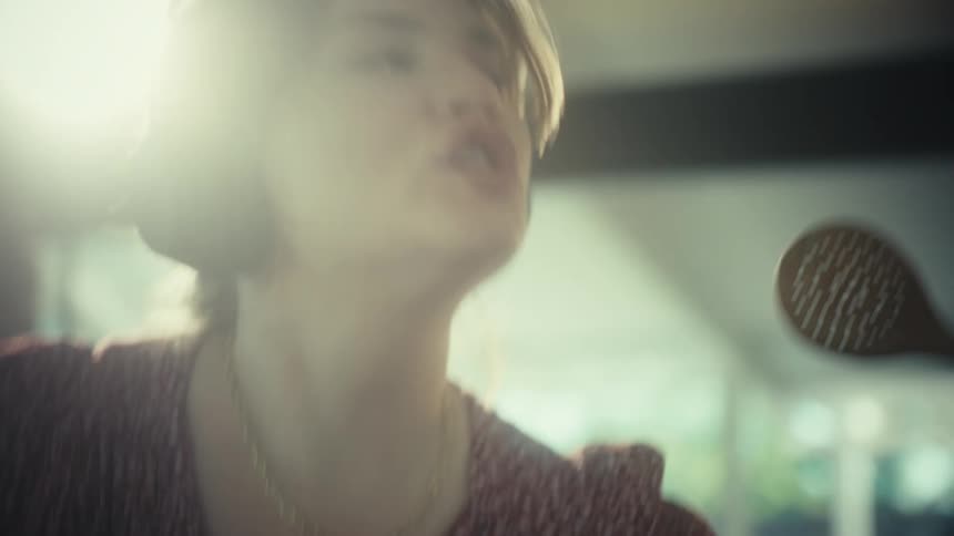 A young woman wearing large headphones sings loudly and dances while using a wooden spoon as a microphone in a kitchen