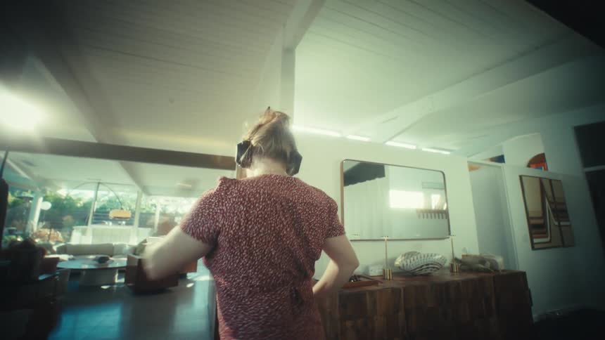 A woman with headphones dances in a sunlit modern living room while wearing a red patterned dress near a wooden sideboard and a brick fireplace