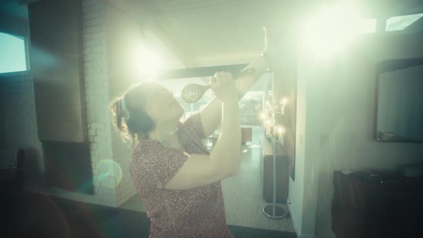 A young woman wearing large headphones sings loudly and dances while using a wooden spoon as a microphone in a kitchen