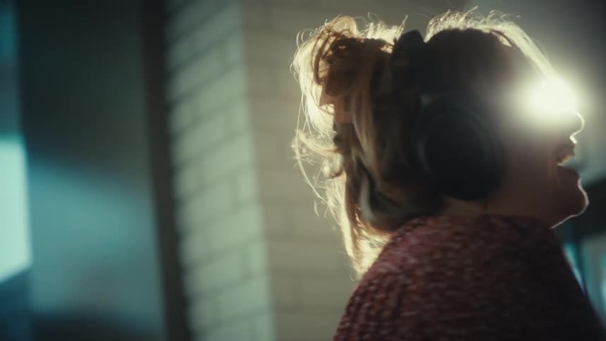 A woman wearing large headphones dances and sings joyfully into a wooden laddle her laughter and energetic movements filling a sundrenched room as bright light flares from a nearby window