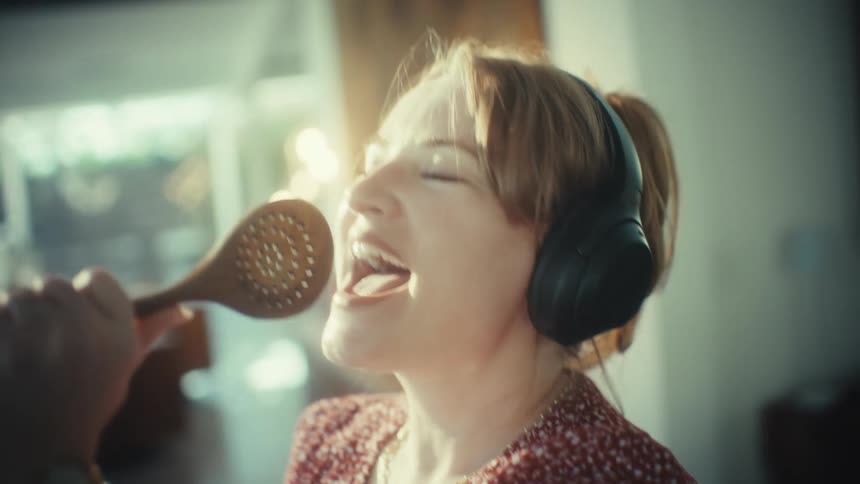 A young woman in her living room wearing a headset holding a spatula like a microphone as she sings and has fun