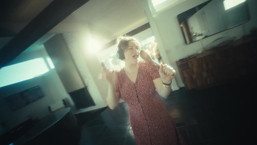 A young woman wearing large headphones sings loudly and dances while using a wooden spoon as a microphone in a kitchen