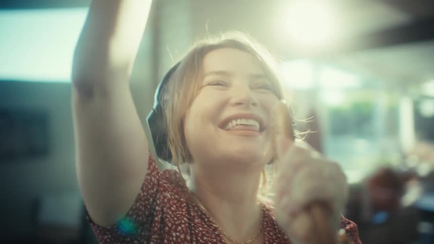 A young woman wearing large headphones sings loudly and dances while using a wooden spoon as a microphone in a kitchen