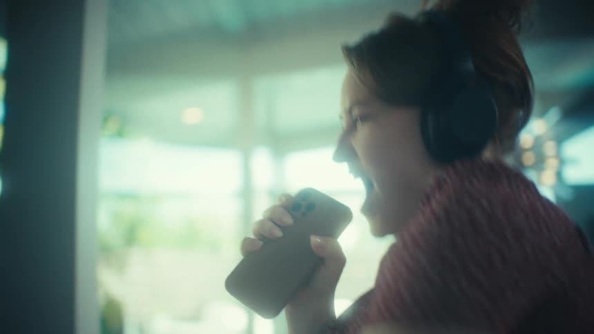 A young woman wearing large headphones sings loudly and dances while using a smartphone as a microphone in a kitchen