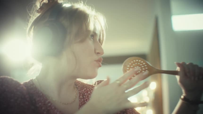 A young woman wearing large headphones sings loudly and dances while using a wooden spoon as a microphone in a kitchen
