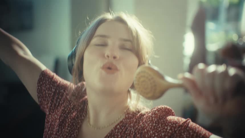 In a sunlit living room a woman wearing headphones sings passionately into a wooden ladle her face lit with a bright smile as she dances and enjoys a carefree musical moment