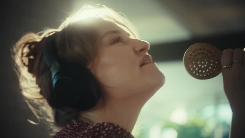 A young woman wearing large headphones sings loudly and dances while using a wooden spoon as a microphone in a kitchen