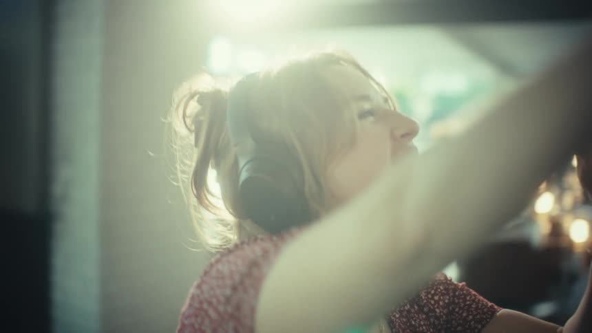 A young woman wearing large headphones sings loudly and dances while using a wooden spoon as a microphone in a kitchen