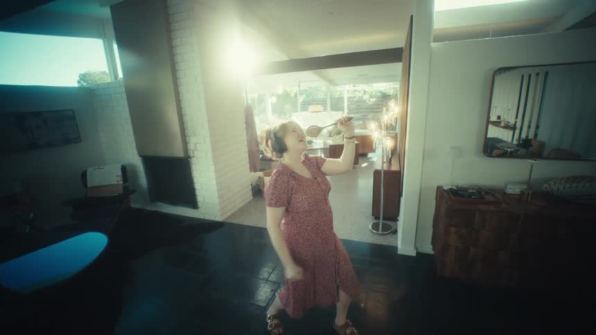 A woman wearing headphones sings into a kitchen utensil while dancing in a living room