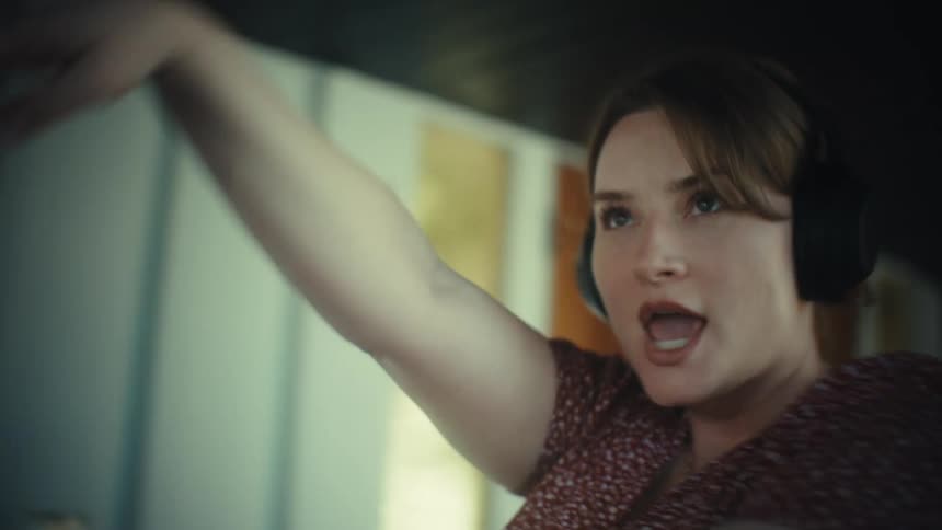 A young woman wearing large headphones sings loudly and dances while holding a phone like a microphone in a kitchen