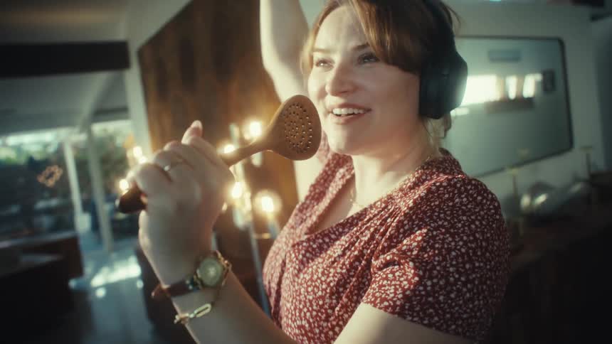 A young woman wearing large headphones sings loudly and dances while using a wooden spoon as a microphone in a kitchen