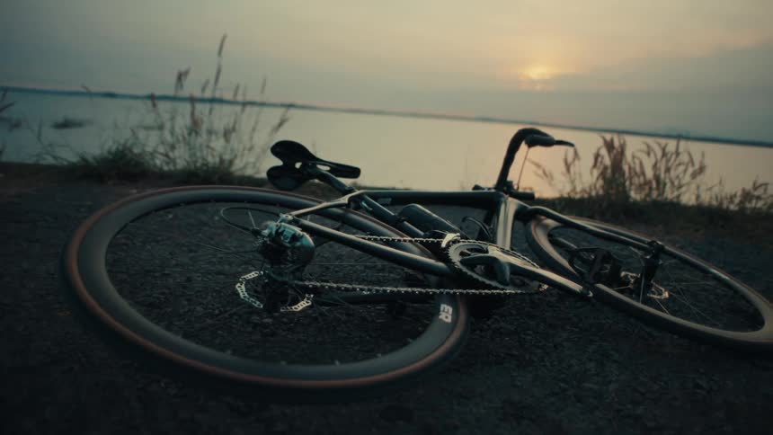 A black road bicycle lies on a gravel path against the backdrop of a calm lake at sunset