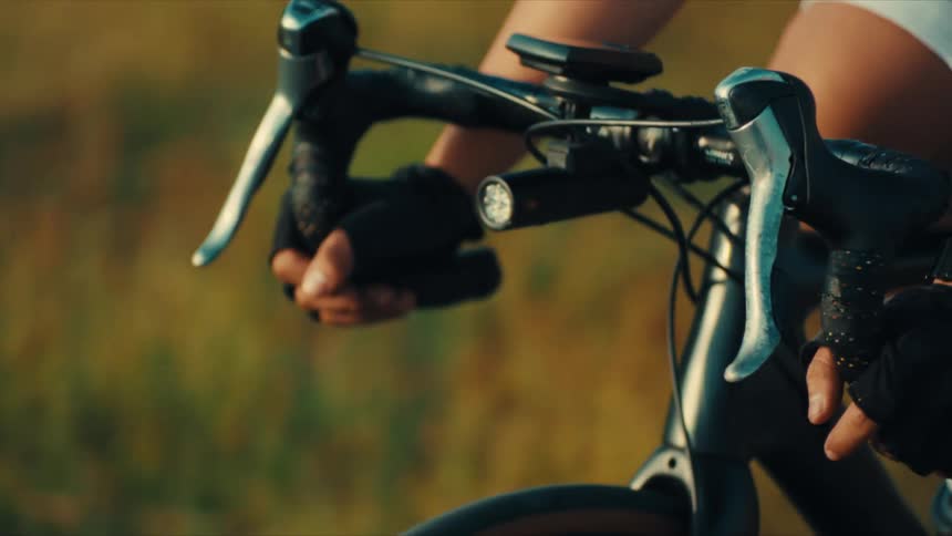 A close view shows the gloved hands of a cyclist gripping the drop handlebars of a road bike while riding past a field at sunset