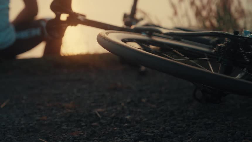 A cyclist sitting by a body of water at sunset with their road bike resting on the gravelly ground in the foreground the camera slowly pans across the bicycles rear wheel and frame capturing the warm golden light reflecting off the spokes and the wat
