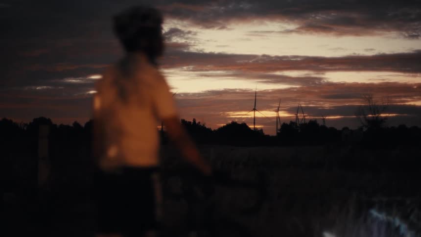 A view from behind shows a cyclist standing next to a road bike during a dark sunset
