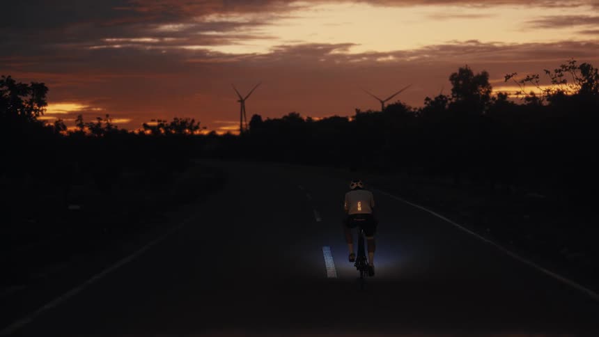 A cyclist rides a bicycle down a rural road at sunset with wind turbines in the background