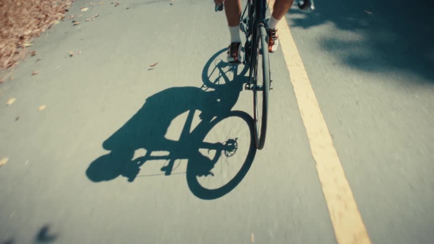 A cyclist pedals steadily along a sundrenched road their dark shadow stretching across the asphalt as they pass over scattered dry leaves and follow the path of a weathered yellow line
