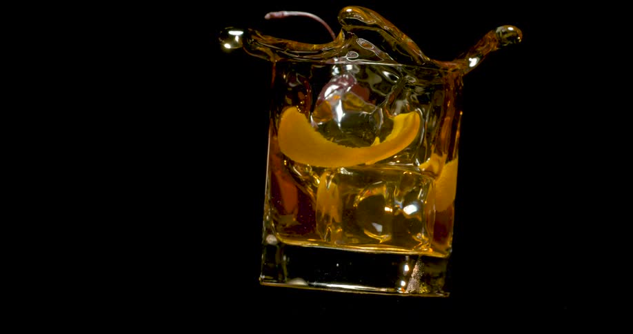 An amber cocktail with a cherry and orange peel splashes out of a crystal glass against a black background in slow motion