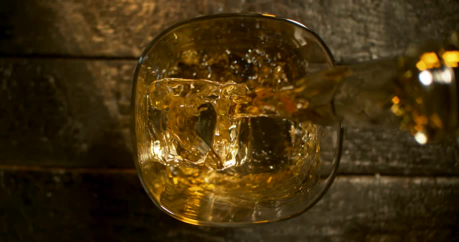 Amber liquor pours from a bottle onto the ice cubes of a glass sitting on a wooden surface