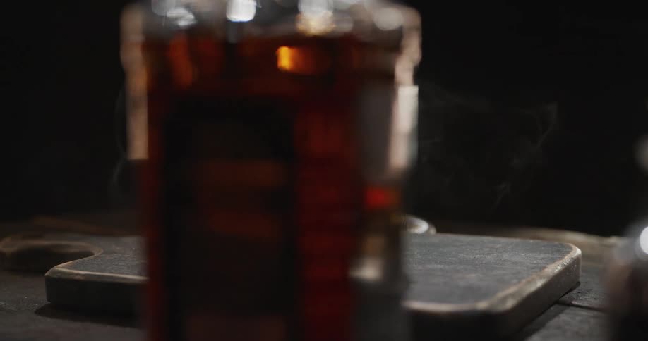 A view of a smoky glass of whiskey on a dark stone board shows white smoke curling from the ice as a camera moves around the scene in a circle