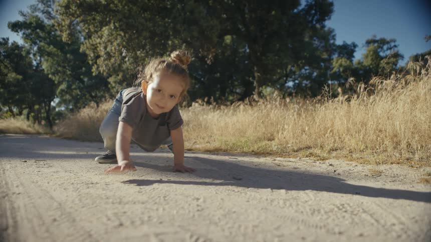 A young child with a bun crawls forward on a dirt path in a sunny field the toddler smiles at the camera while surrounded by tall dry grass and trees