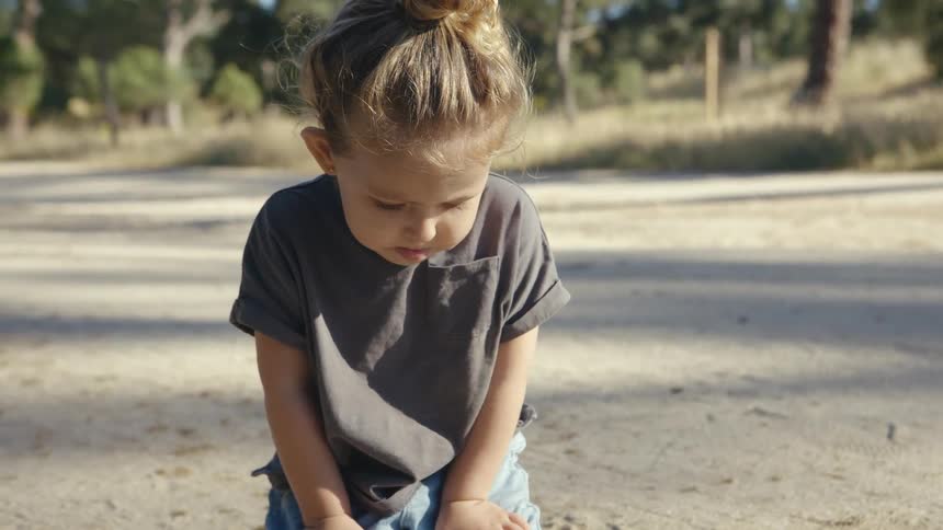 A young child with hair tied in a bun sits on a sandy path outdoors the child is wearing a grey tshirt and appears focused while playing on the ground