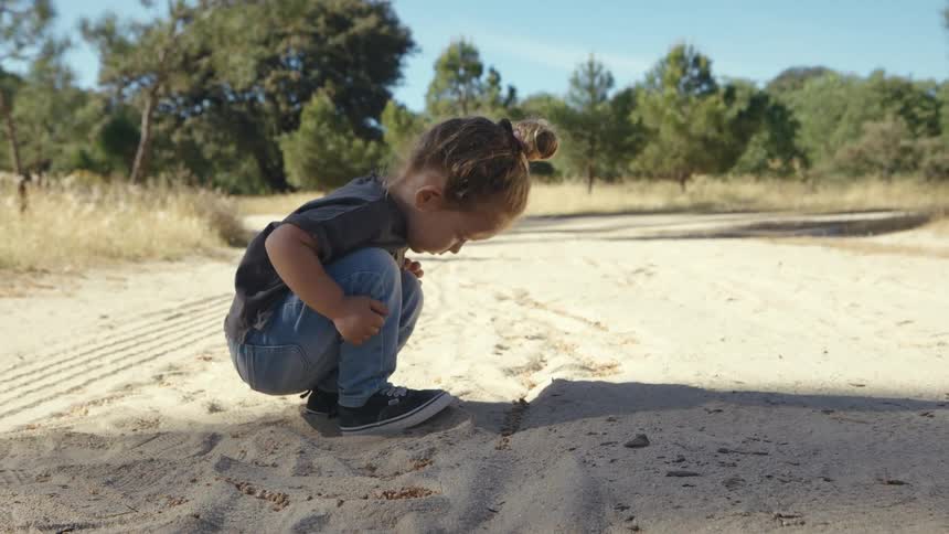 Little girl crouches while observing marks in the sand on a dirt road surrounded by trees