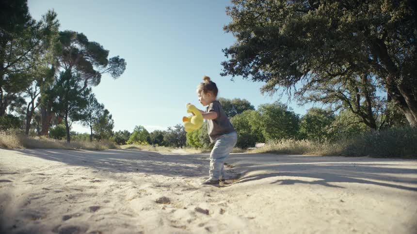A young child carrying a yellow stuffed toy walks and jumps along a wide sandy path through a park filled with green trees under a bright blue sky