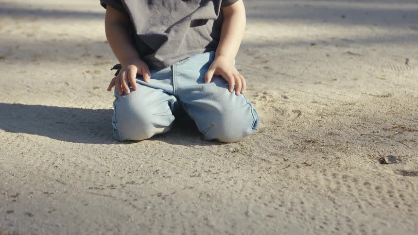 A young child with hair in a bun kneels on a sandy path in a park while looking down at the ground