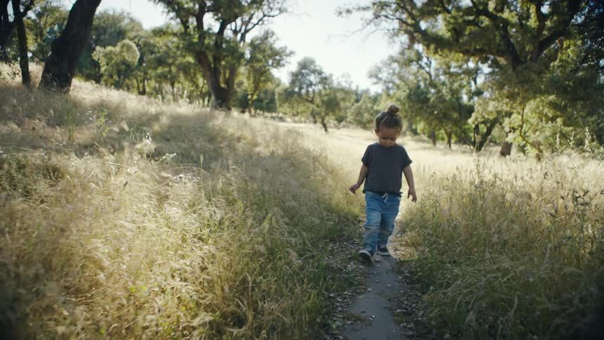 A young child with hair in a bun walks slowly along a narrow dirt path through a field of tall dry grass and green trees