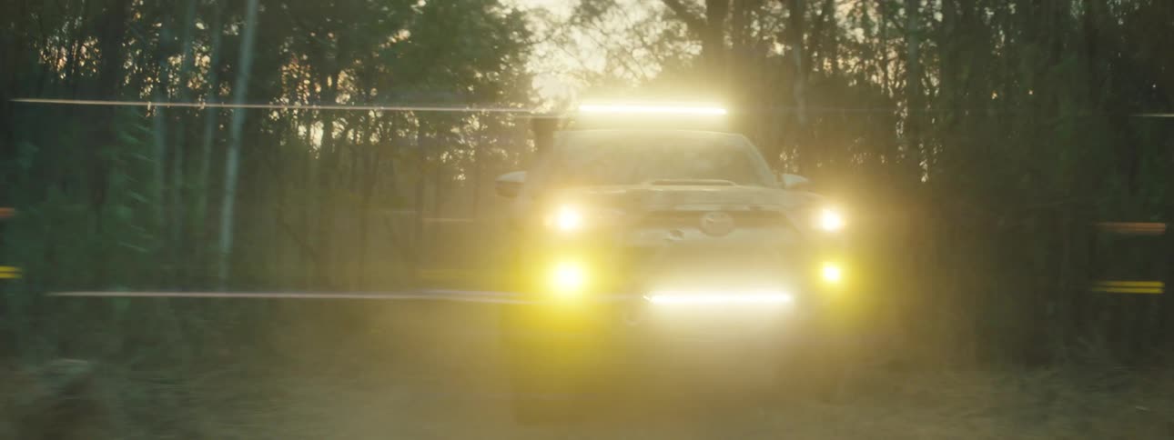 A vehicle with bright headlights drives along a dirt road through a forest at sunset