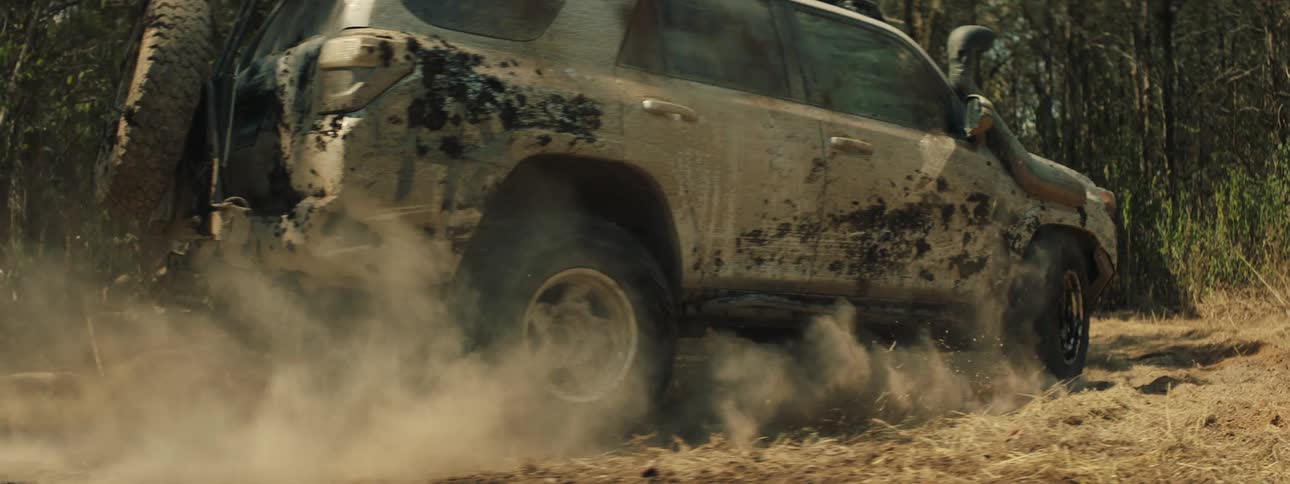 A tracking shot shows a tan vehicle covered in mud as it drives quickly through a wooded area while kicking up a thick cloud of dust from the ground