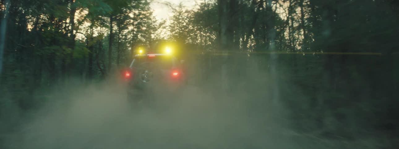 A dirty off road suv speeds through a dense forest on a dusty trail during sunset while its roof lights shine and the wheels kick up a large cloud of brown earth in a motion blur