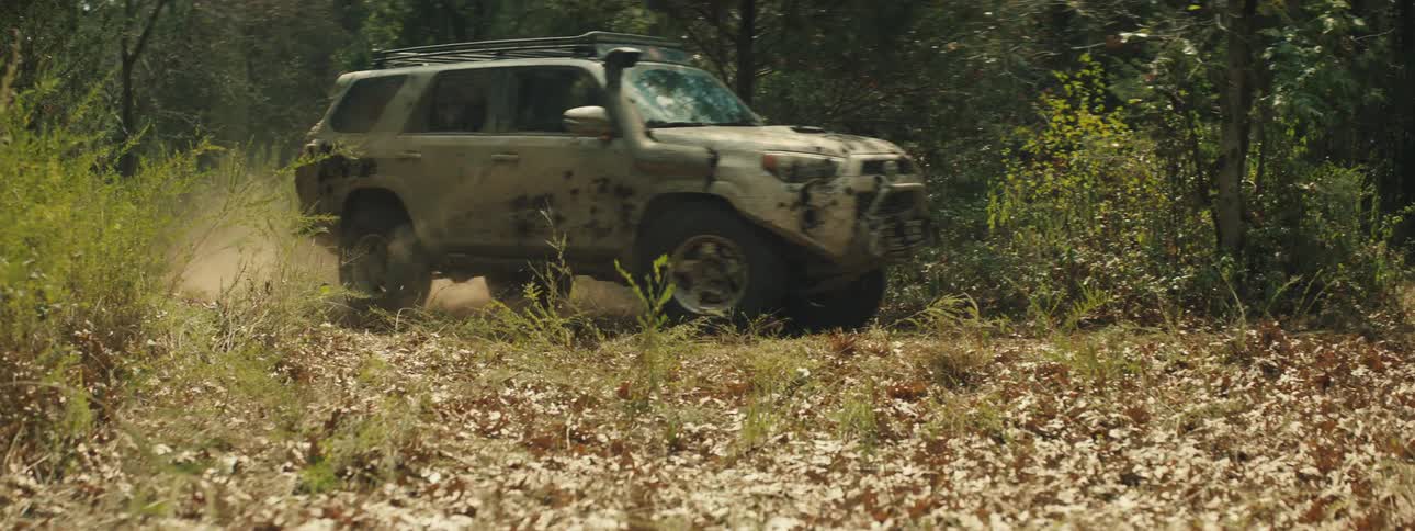 A tan off road vehicle covered in mud drives quickly across a field of dry leaves and kicks up a large cloud of dust